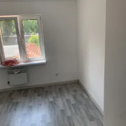 Pomalowane na biało wnętrze pokoju z szarą podłogą imitującą drewno, grzejnikiem elektrycznym pod oknem i zwiniętym przewodem na parapecie.