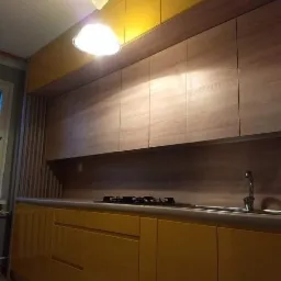 Żółto-drewniana zabudowa kuchenna z górnymi szafkami pod sufitem i oświetleniem podszafkowym, widoczny zlew i płyta gazowa.