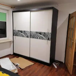 Wnętrze pokoju z dużą szafą przesuwną w kolorach bieli i grafitu, z poziomym pasem dekoracyjnym w motywie zebry. Widoczne ślady montażu: kartony i akcesoria na podłodze.