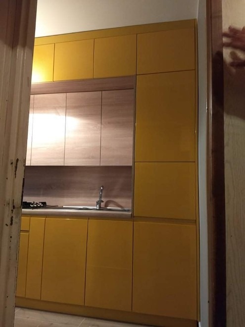 Żółta zabudowa kuchenna z drewnianymi frontami szafek wiszących i blatem, widok zza framugi drzwi.