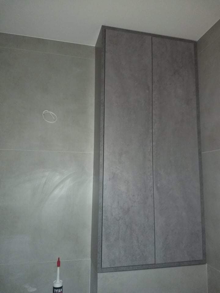 Szara, dwudrzwiowa szafka wnękowa z fakturą betonu, zamontowana w łazience wykończonej szarymi płytkami, widoczna tuba silikonu montażowego u dołu kadru, Pionki.
