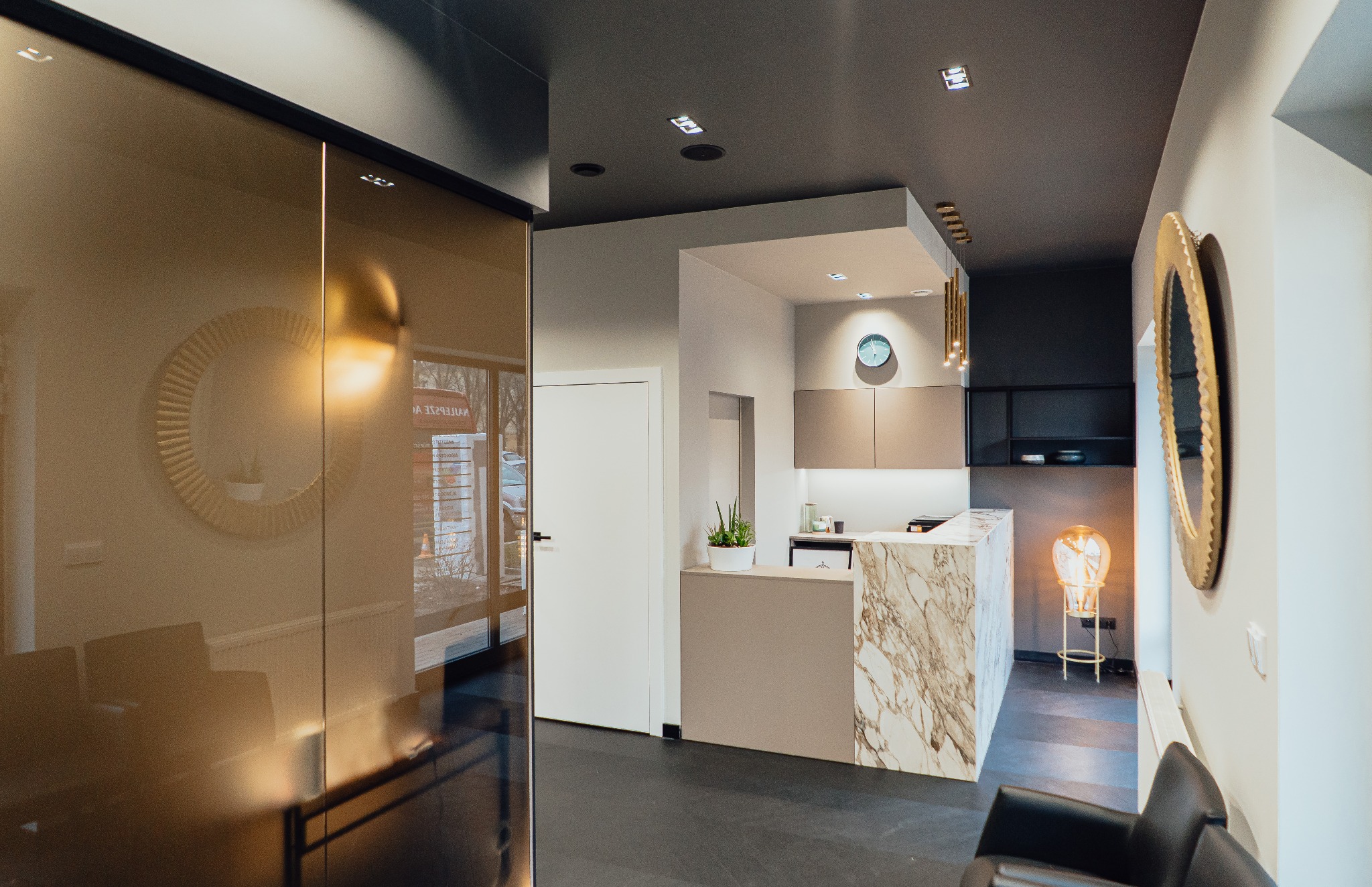 Recepcja gabinetu stomatologicznego z marmurowym blatem, widok zza szklanych drzwi z odbiciem okrągłego lustra, nowoczesne oświetlenie i minimalistyczny design.