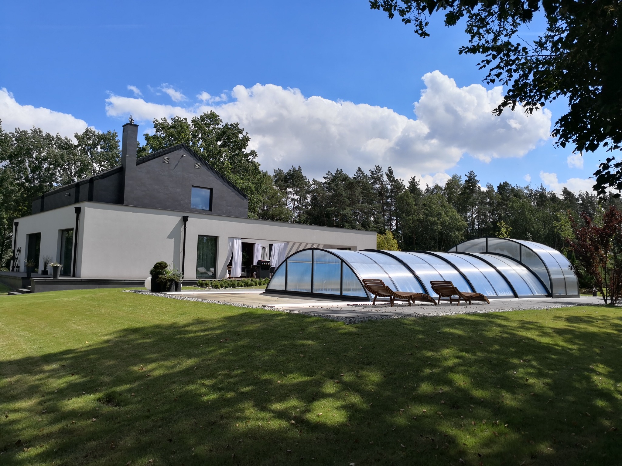 Nowoczesny dom jednorodzinny z płaskim dachem, szarą elewacją i przeszkloną zabudową basenową z leżakami, otoczony zielonym trawnikiem i drzewami w słoneczny dzień.