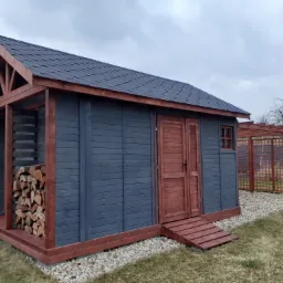 Drewniana konstrukcja z zadaszeniem, pomalowana na szaro z brązowymi elementami wykończeniowymi, służąca jako składzik na drewno i narzędzia ogrodowe, z widocznym ułożonym drewnem i małym oknem...