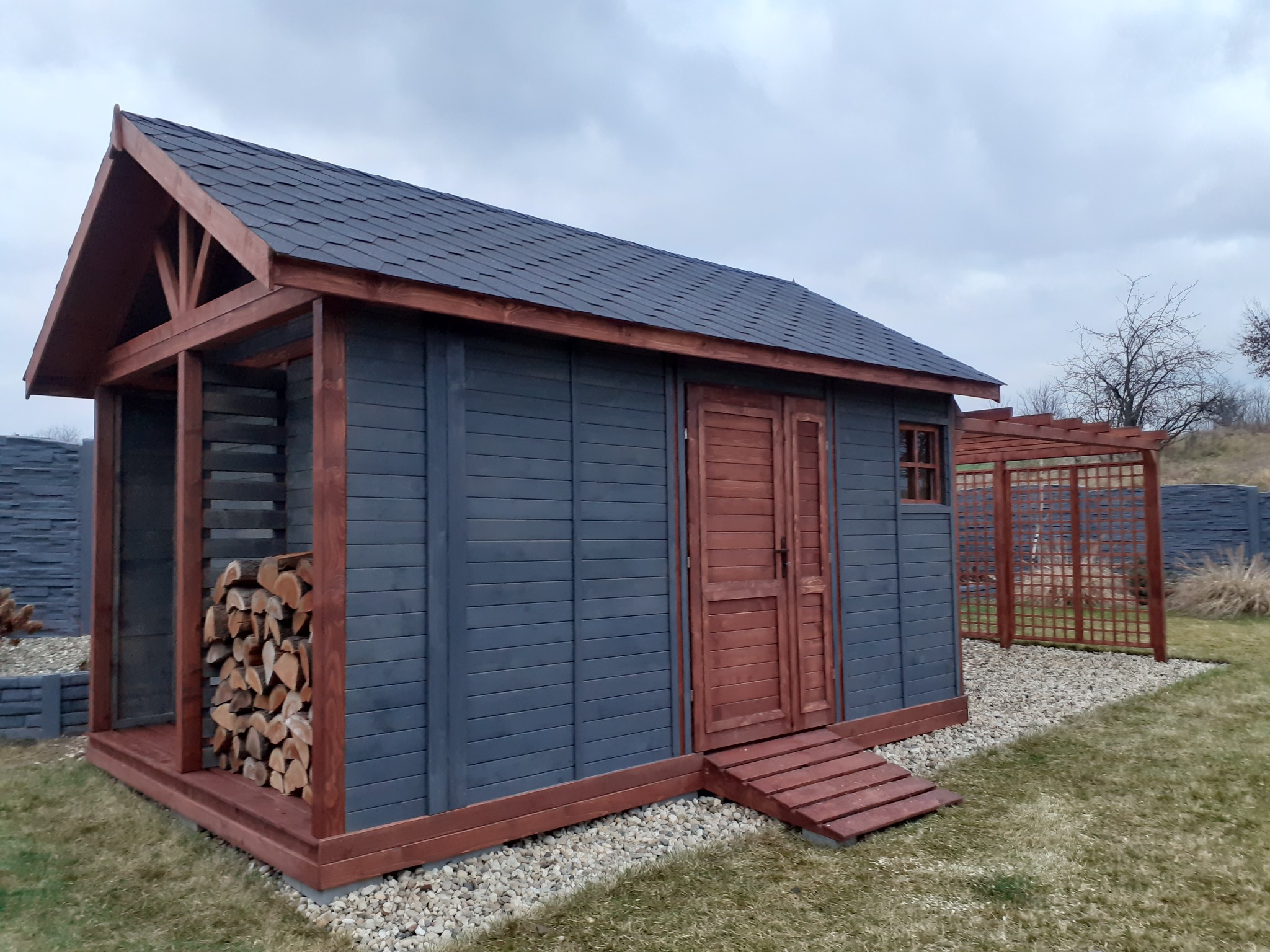 Drewniana konstrukcja z zadaszeniem, pomalowana na szaro z brązowymi elementami wykończeniowymi, służąca jako składzik na drewno i narzędzia ogrodowe, z widocznym ułożonym drewnem i małym oknem...