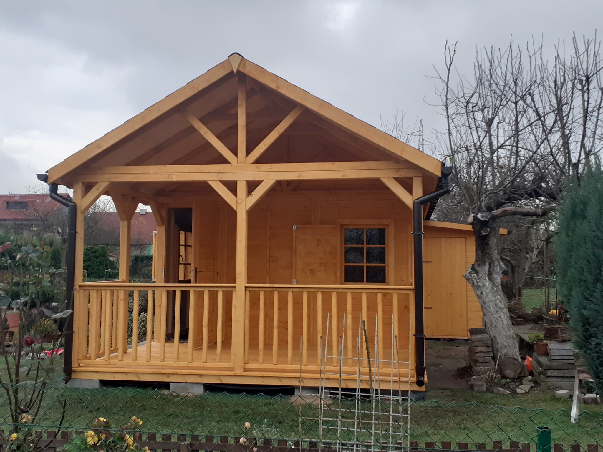 Nowa, surowa, drewniana konstrukcja z zadaszonym tarasem, widoczna z zewnątrz, z elementami wykończeniowymi takimi jak rynny, na tle pochmurnego nieba i jesiennego ogrodu.