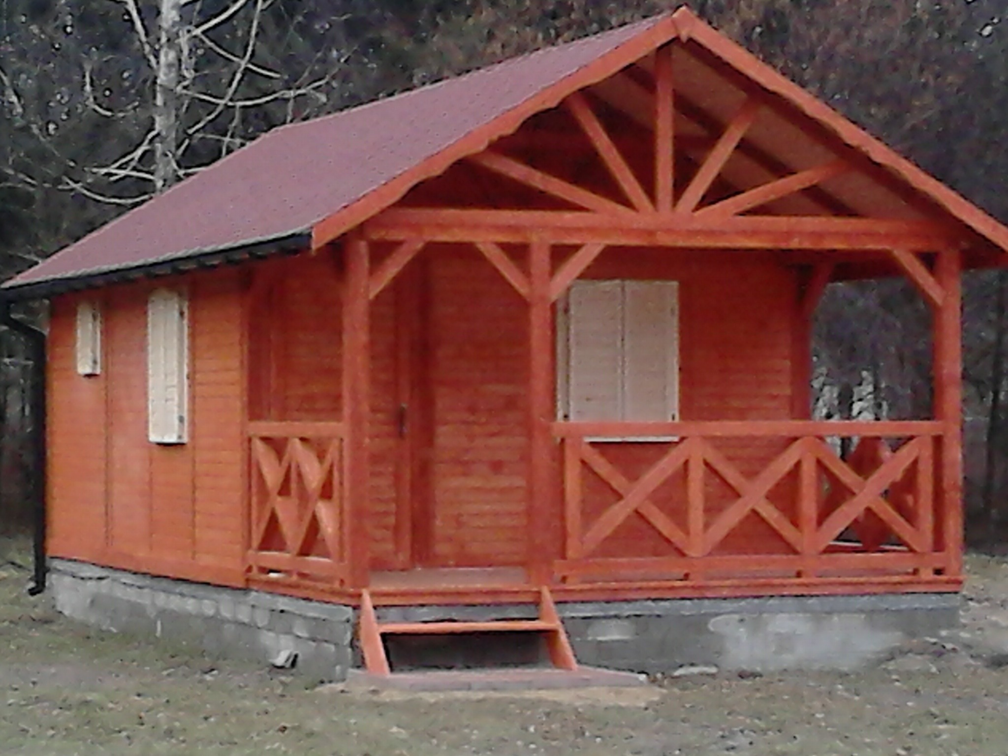 Drewniana konstrukcja z zadaszeniem i balustradą w kolorze pomarańczowym, z dwoma oknami z okiennicami, na betonowej podstawie, w tle drzewa.