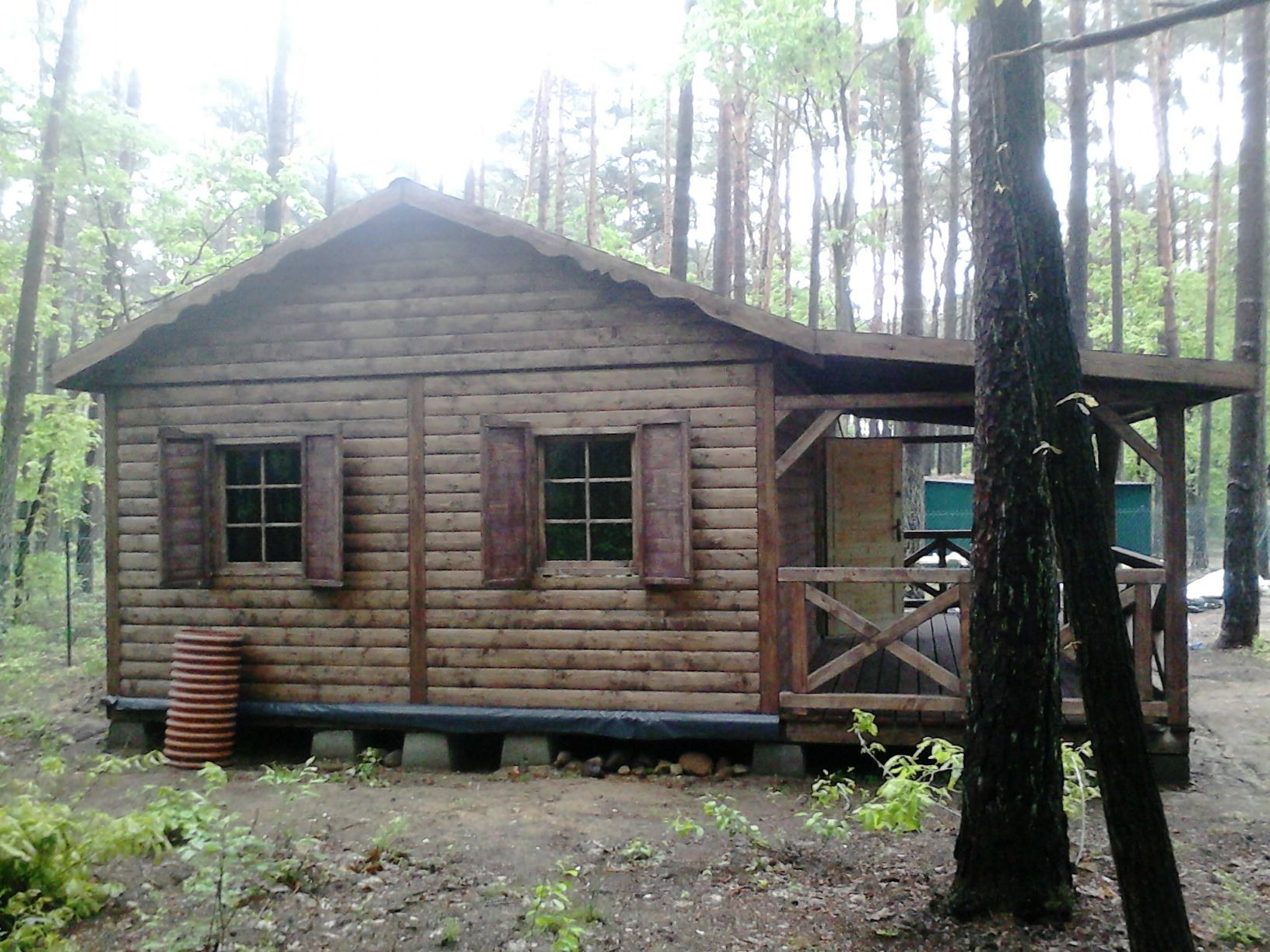 Drewniana konstrukcja w lesie z zadaszonym tarasem, okna z okiennicami, widoczne podpory betonowe i ułożone rury drenarskie.