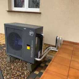 Szara jednostka zewnętrzna pompy ciepła EcoHeat Mono, ustawiona na betonowych podstawach obok budynku z beżową elewacją, widoczne rury w srebrnej izolacji, okno z firanką w tle.