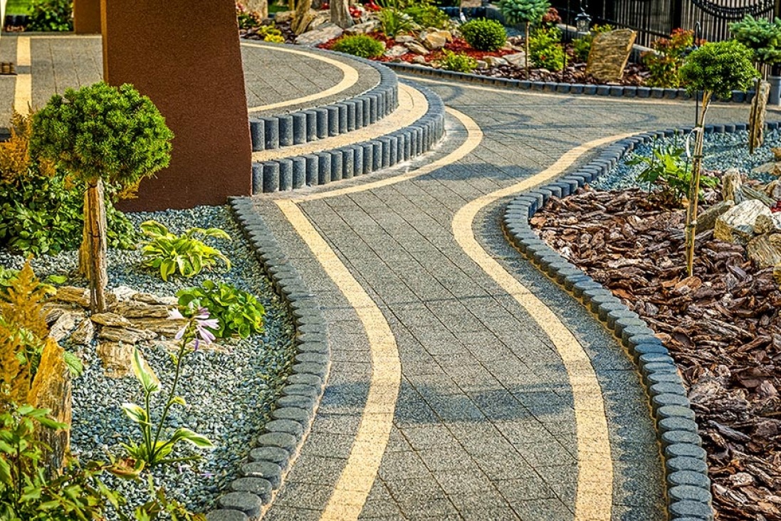 Kręta ścieżka z szarej kostki brukowej z żółtymi pasami, obrzeża z ciemnych, okrągłych elementów, ozdobne rośliny, kamienie i kora wzdłuż ścieżki.