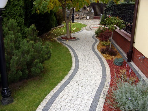 Kręta ścieżka z jasnej kostki granitowej, obramowana ciemniejszą kostką, prowadząca przez zadbany ogród z drzewami i krzewami, widok z góry.