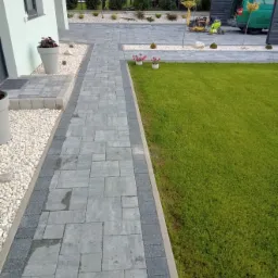 Ścieżka z szarej kostki brukowej z ciemniejszym obramowaniem prowadząca do domu, obok trawnik i rabaty z białym kamieniem, w tle zaparkowany zielony samochód i pracownik z zagęszczarką.