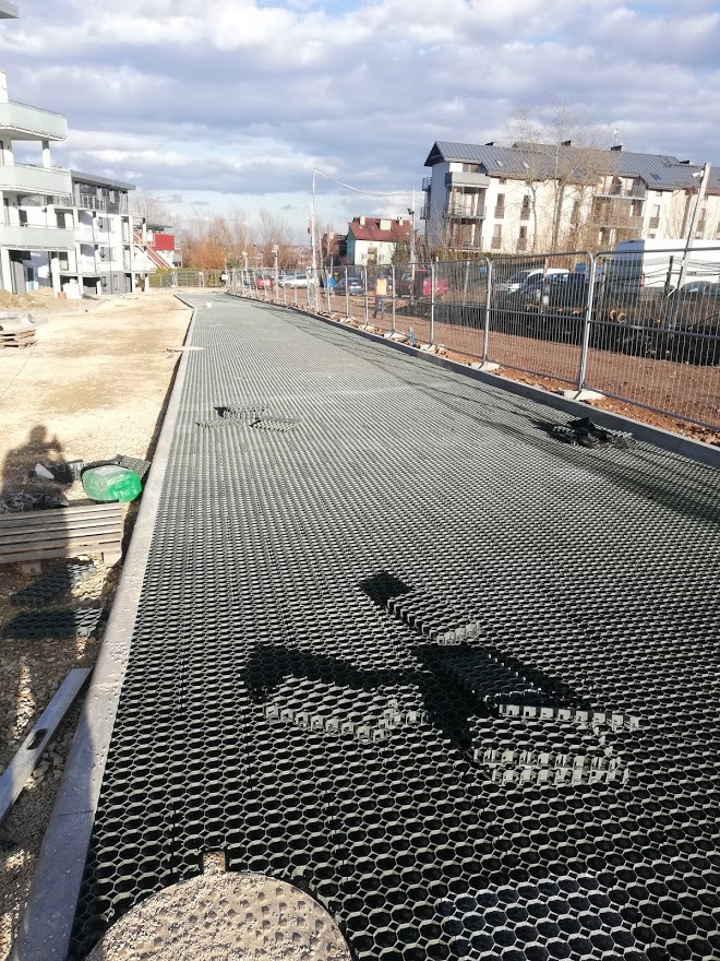 Ułożona, pusta, plastikowa siatka brukowa na podbudowie drogi dojazdowej, widoczne budynki w tle i cień osoby robiącej zdjęcie.