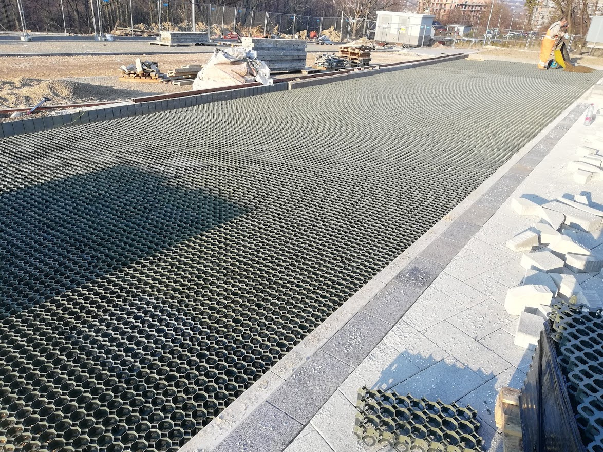 Szeroki kadr na ułożoną, ażurową nawierzchnię z tworzywa sztucznego w kształcie plastra miodu, obok której widać paletę z kolejnymi elementami oraz częściowo ułożoną kostkę brukową po prawej...