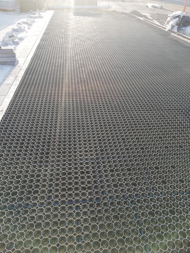 Plastikowa siatka o strukturze plastra miodu przygotowana pod nawierzchnię parkingu lub drogi dojazdowej, z widocznym podsypem piaskowym w oczkach i paletą kostki brukowej w tle.
