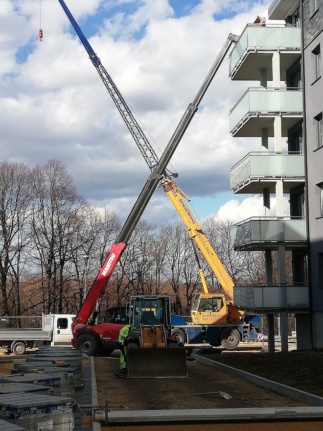 Dwa dźwigi teleskopowe, czerwony Manitou i żółty Dźwig Trans, krzyżują się na tle budynku z balkonami i pochmurnego nieba, obok zaparkowany biały dostawczak i palety kostki brukowej Bruk-Bet...