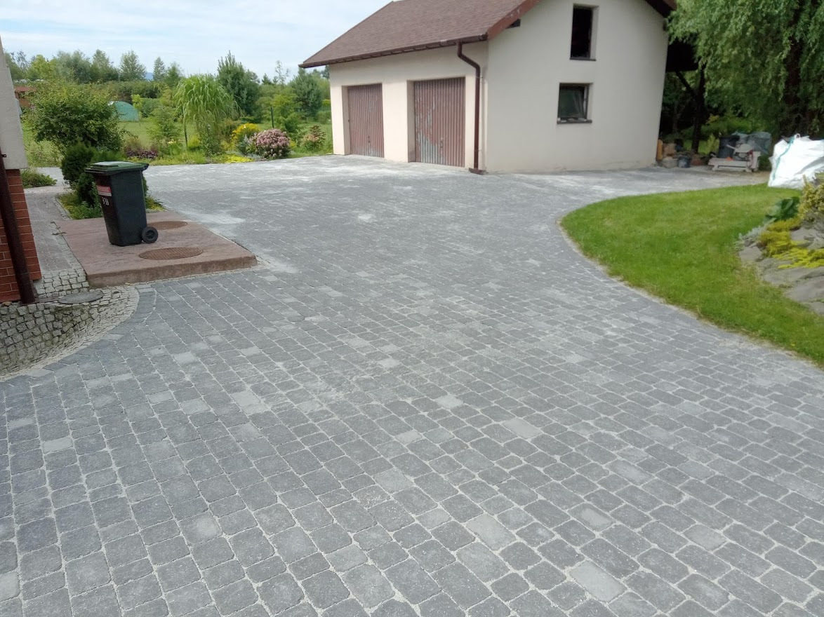 Szeroki podjazd z szarej kostki brukowej prowadzący do garażu z brązowymi drzwiami, obok zielony trawnik i kosz na śmieci.