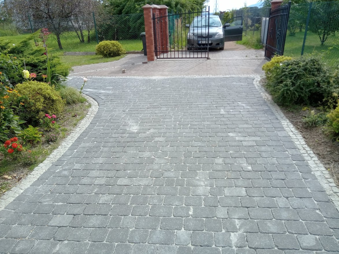 Szeroki podjazd z szarej kostki brukowej prowadzący do bramy z czarnego metalu, obrzeża wykończone jaśniejszą kostką, po bokach rabaty z kwiatami i krzewami, w tle zaparkowany samochód osobowy...