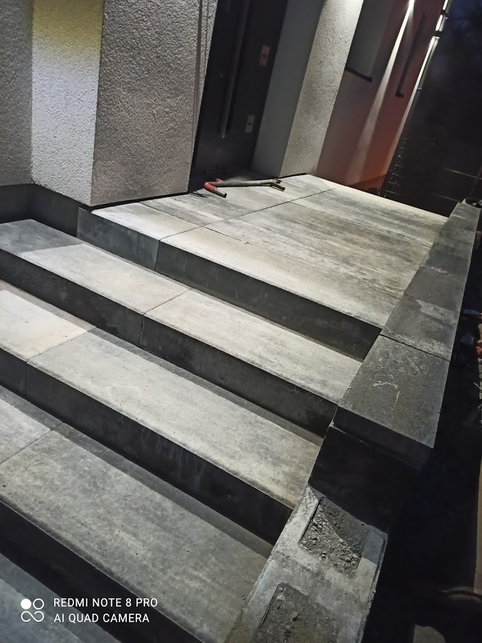 Schody wejściowe wyłożone szarą kostką brukową, widoczne narzędzia budowlane na górnym podeście, oświetlenie zewnętrzne.