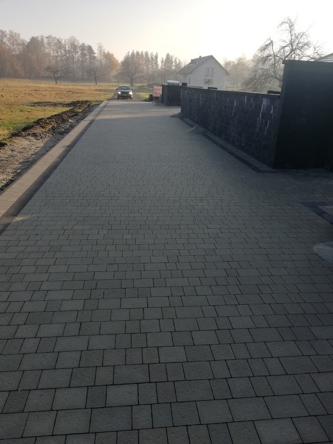 Długa, szara droga z kostki brukowej prowadząca do domu, z zaparkowanym samochodem w oddali, widok z niskiej perspektywy w pochmurny dzień.