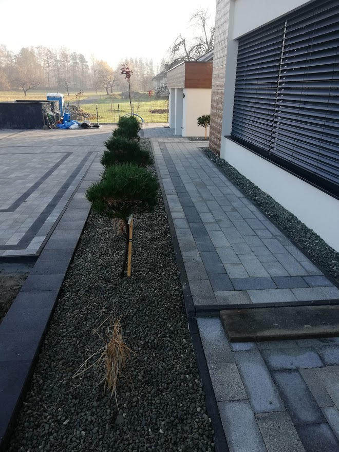 Ułożona szara kostka brukowa z ciemniejszymi pasami, oddzielona rabatą z kamykami i trzema młodymi drzewkami bonsai, widok na dom z żaluzjami i oddalony krajobraz.