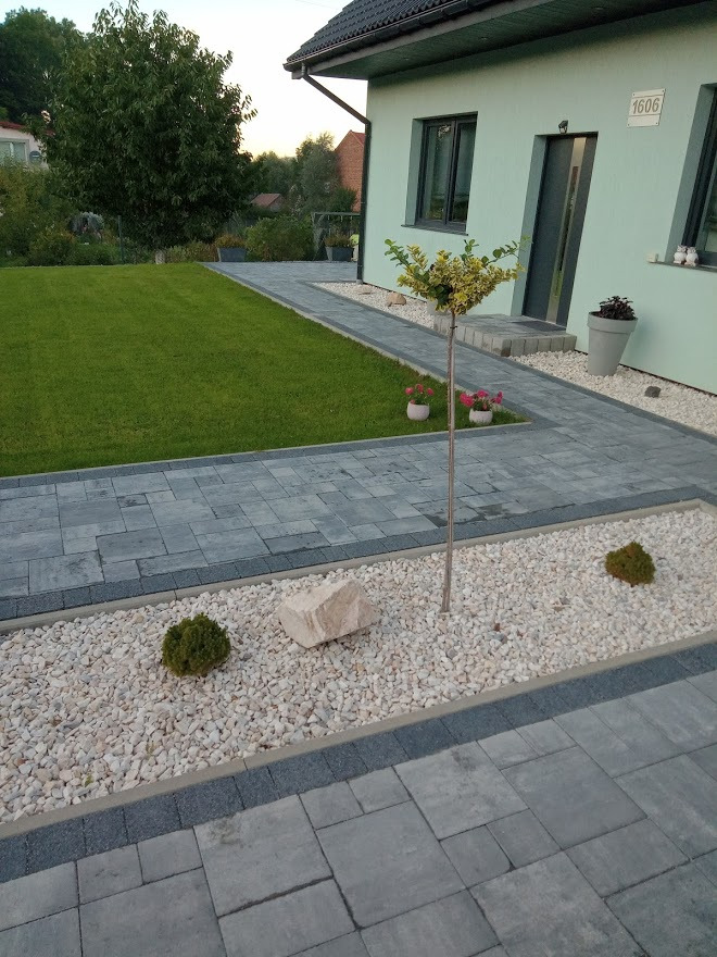 Nowoczesny podjazd z szarej kostki brukowej prowadzący do jasnego domu z numerem 1606, ozdobiony białymi kamykami, pojedynczym głazem i niskimi krzewami.
