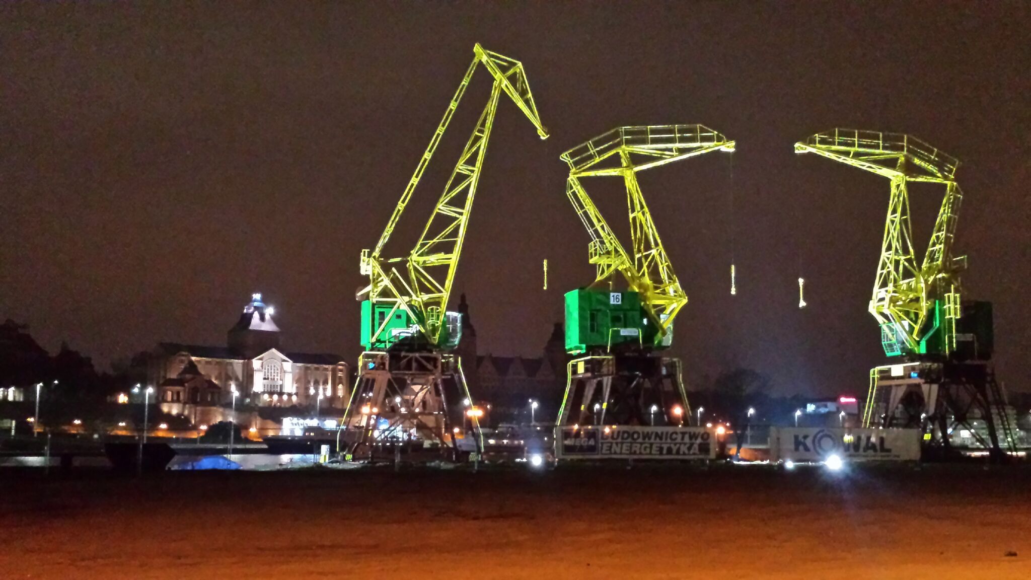 Nocny widok na trzy podświetlone na zielono i żółto dźwigi portowe, z budynkiem w tle i oświetleniem miejskim.