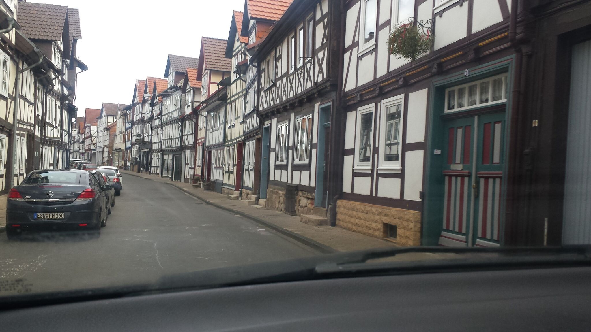 Ulica z rzędem budynków w stylu szachulcowym, widok zza szyby samochodu, zaparkowane auta po lewej stronie.