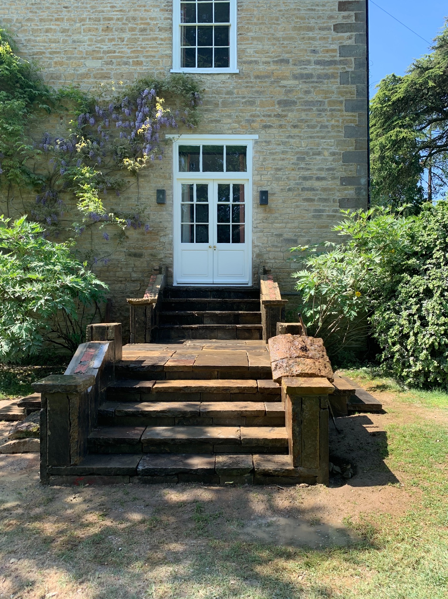 Kamienny budynek z białymi drzwiami i oknem, porośnięty bluszczem z fioletowymi kwiatami, z kamiennymi schodami wejściowymi.
