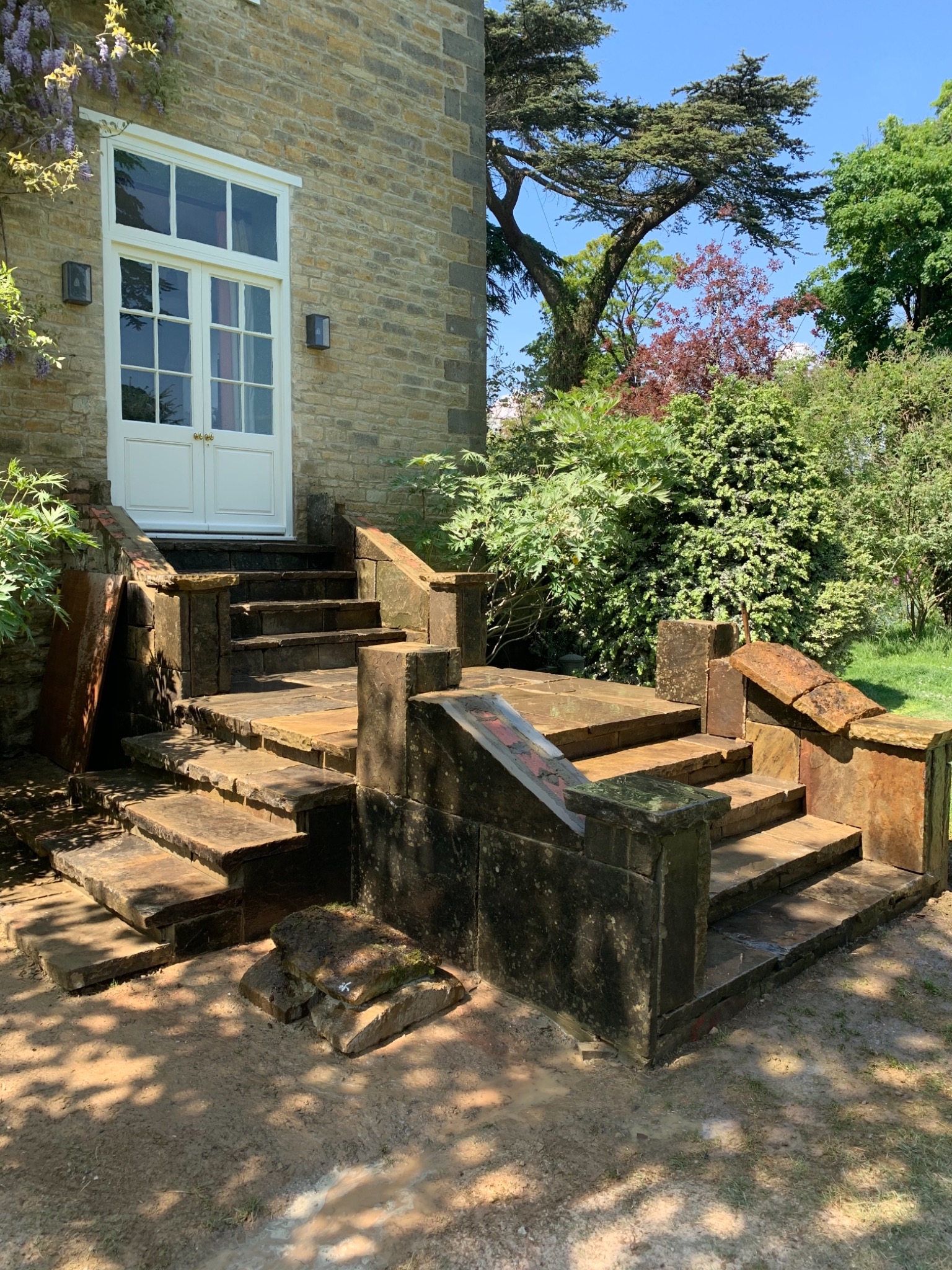 Kamienne schody wejściowe do domu z białymi drzwiami, otoczone zielenią, z widocznymi śladami prac remontowych i luźnymi kamieniami na ziemi.