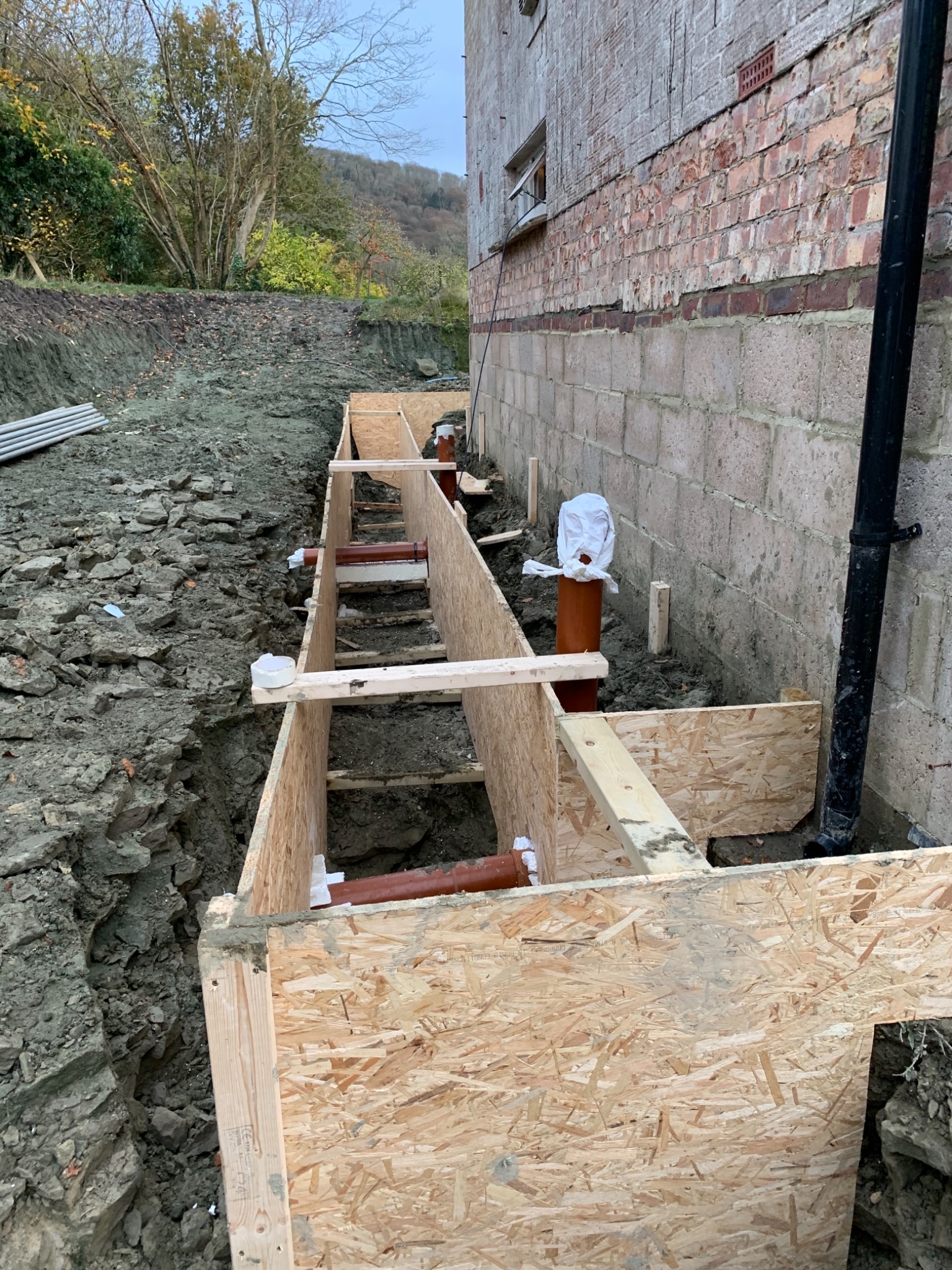 Wykop pod fundamenty budynku z drewnianym szalunkiem, widoczne rury kanalizacyjne i fragment muru z cegły