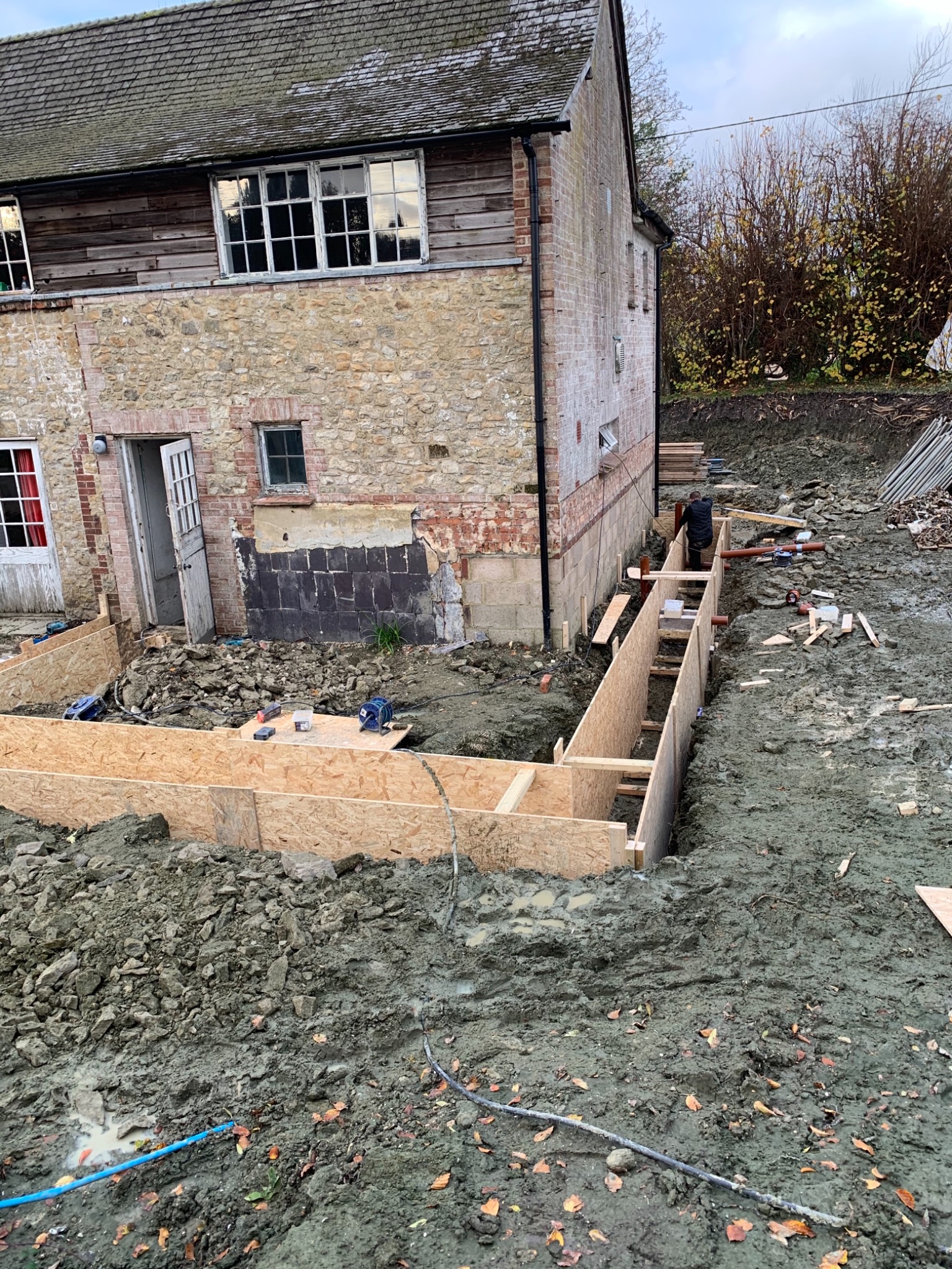 Fundamenty budynku w trakcie budowy: drewniane szalunki w wykopie, widoczne fragmenty muru z cegły i kamienia, teren wokół pokryty błotem i gruzem, w tle elewacja budynku z oknami.