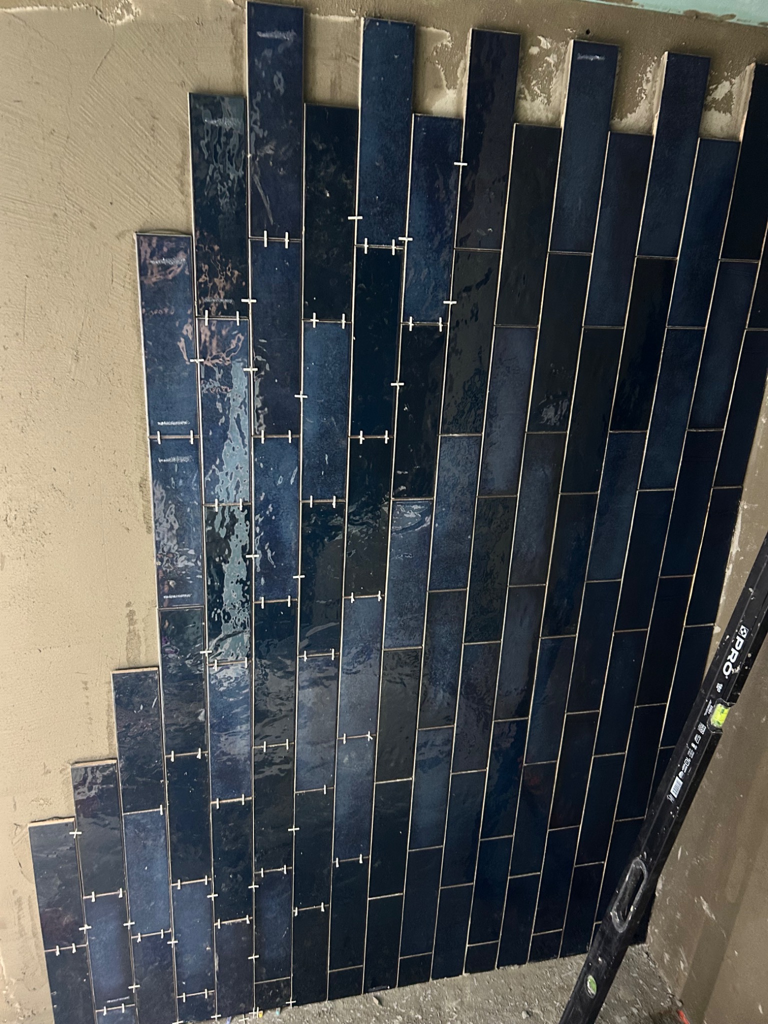 Układanie pionowych, błyszczących, ciemnoniebieskich płytek w kształcie cegiełek na ścianie, widoczne plastikowe krzyżyki dystansowe i poziomica przy ścianie.