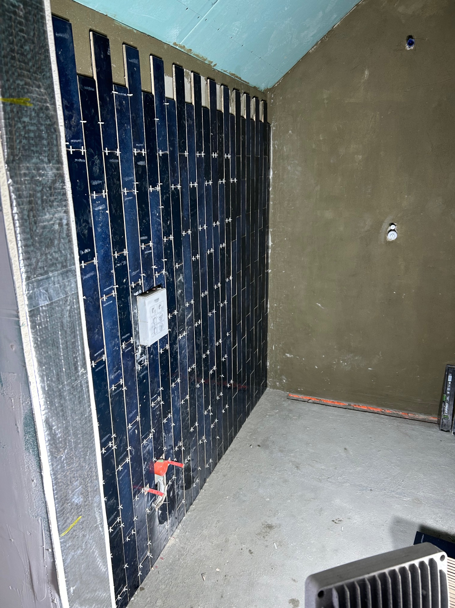 Ściana w trakcie układania pionowych, wąskich, ciemnych płytek, z krzyżykami dystansowymi, widoczna instalacja podtynkowa i surowy tynk na ścianach w tle.