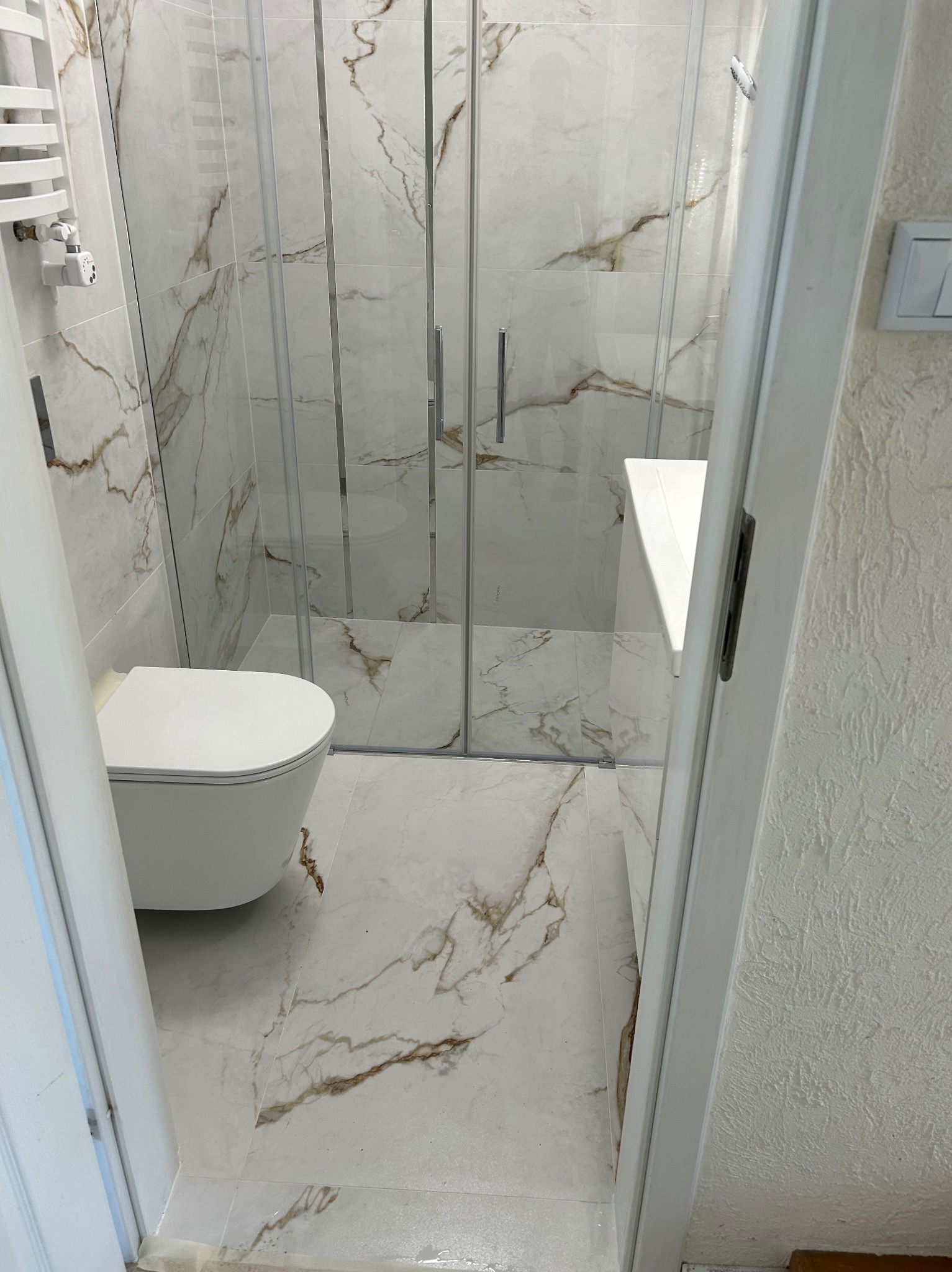 Wykończona łazienka z kabiną prysznicową, toaletą wiszącą i grzejnikiem drabinkowym. Białe płytki imitujące marmur z brązowymi żyłami na ścianach i podłodze. Widoczny fragment ściany z białą...