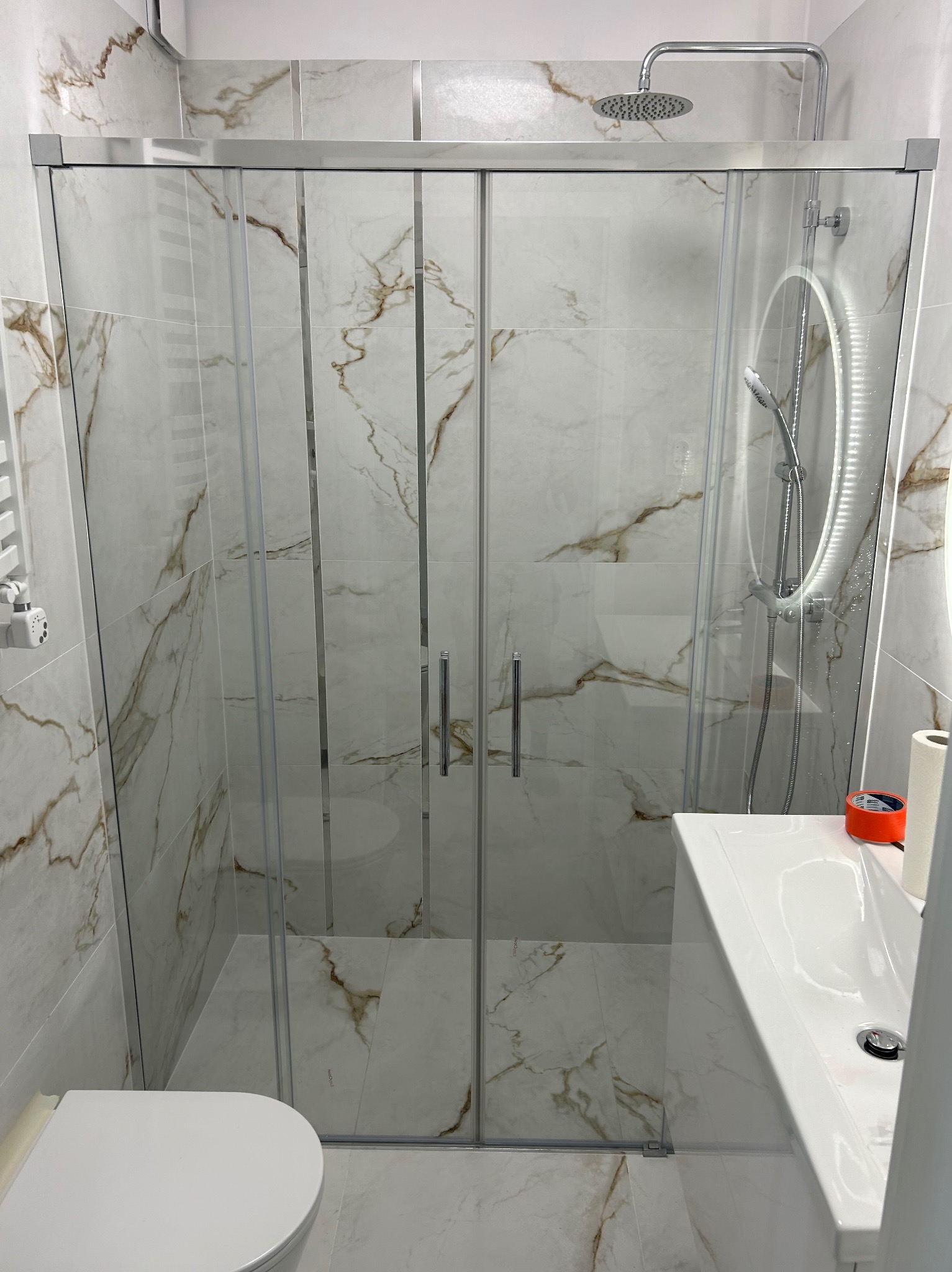 Nowoczesna łazienka z kabiną prysznicową z przesuwnymi drzwiami, wykończona płytkami imitującymi marmur z brązowymi żyłami, widoczna umywalka z oświetlonym lustrem i toaleta.