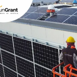 Montaż paneli słonecznych na dachu budynku, pracownik w kasku i kurtce firmowej SunGrant na podnośniku koszowym, widok na zainstalowane panele i elementy wentylacji.