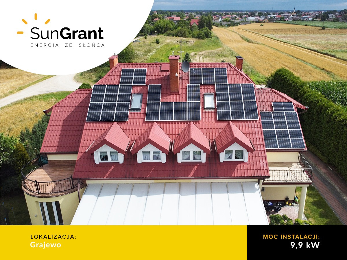 Dom jednorodzinny z czerwoną dachówką, na której zamontowane są panele słoneczne; widok z lotu ptaka, w tle pole i zabudowania.