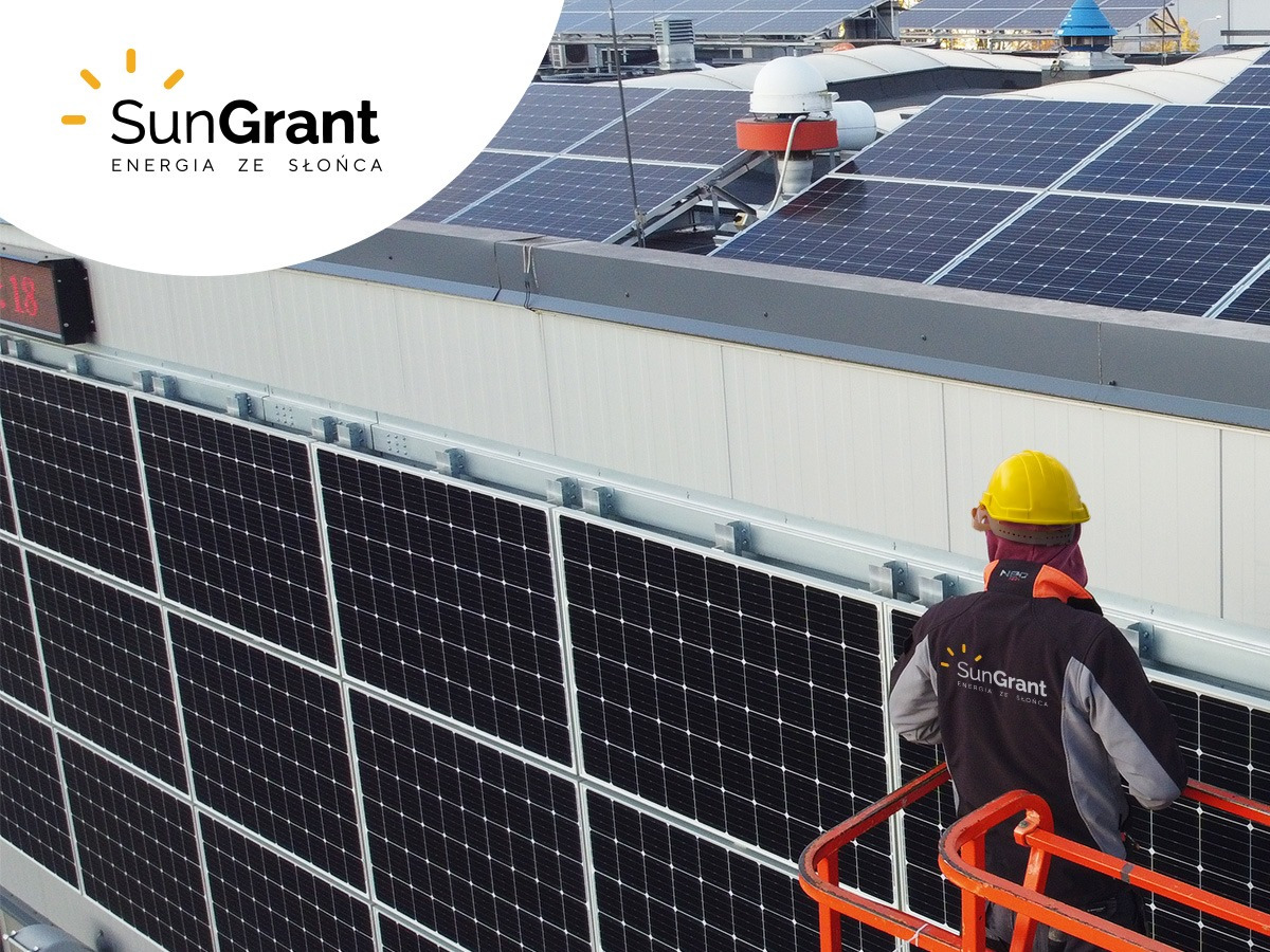 Montaż paneli słonecznych na dachu budynku, pracownik w kasku i kurtce firmowej SunGrant na podnośniku koszowym, widok na zainstalowane panele i elementy wentylacji.