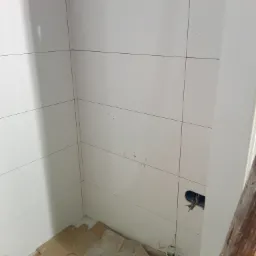 Świeżo ułożone, białe, błyszczące płytki w łazience, widoczne ślady kleju, otwór na instalację elektryczną, zabezpieczone podłoże kartonem.