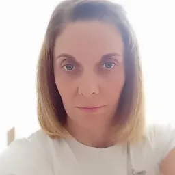 Portret kobiety z blond włosami, ubrana w białą koszulkę z rysunkami, na jasnym tle.