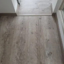 Jasne panele podłogowe z widocznymi słojami drewna, ułożone wzdłuż progu drzwiowego z białą listwą przypodłogową.