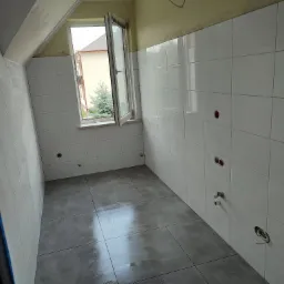 Wnętrze łazienki w Kamieniu po remoncie: białe płytki ścienne z widocznymi otworami na instalacje, szare płytki podłogowe, okno z widokiem na zieleń.