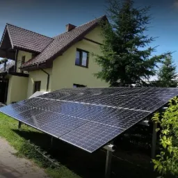 Instalacja fotowoltaiczna na trawniku przed domem jednorodzinnym z brązowym dachem i żółtą elewacją, widok z niskiej perspektywy.