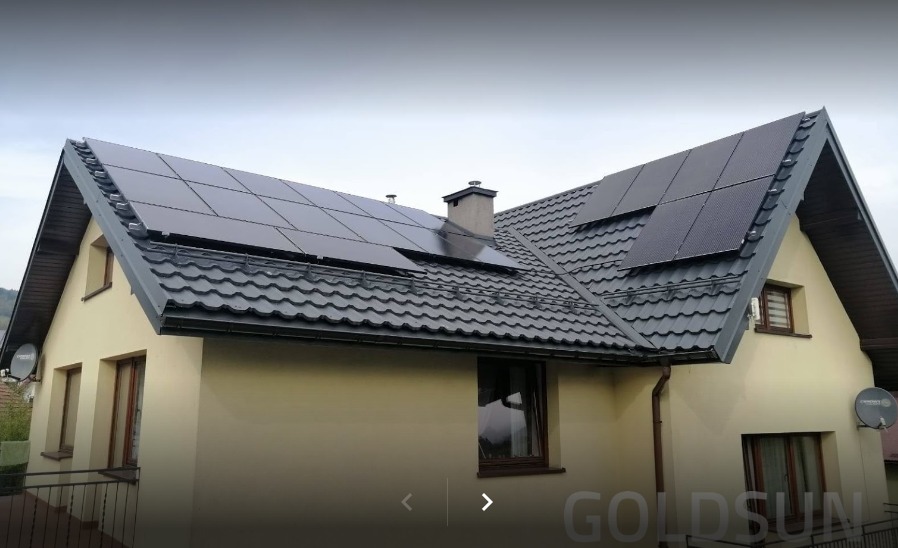 Dom jednorodzinny z zamontowanymi panelami słonecznymi na dachu pokrytym blachodachówką w kolorze grafitowym, widoczne okna i antena satelitarna.