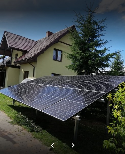 Instalacja fotowoltaiczna na trawniku przed domem jednorodzinnym z brązowym dachem i żółtą elewacją, widok z niskiej perspektywy.