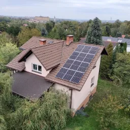 Widok z góry na dom jednorodzinny z brązowym dachem i panelami słonecznymi, otoczony zielenią, w tle widoczny krajobraz miejski.