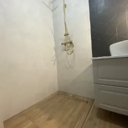 Nowoczesna kabina prysznicowa walk-in z odpływem liniowym w podłodze wyłożonej płytkami imitującymi drewno, złota armatura i czarne płytki z geometrycznym wzorem na ścianie.