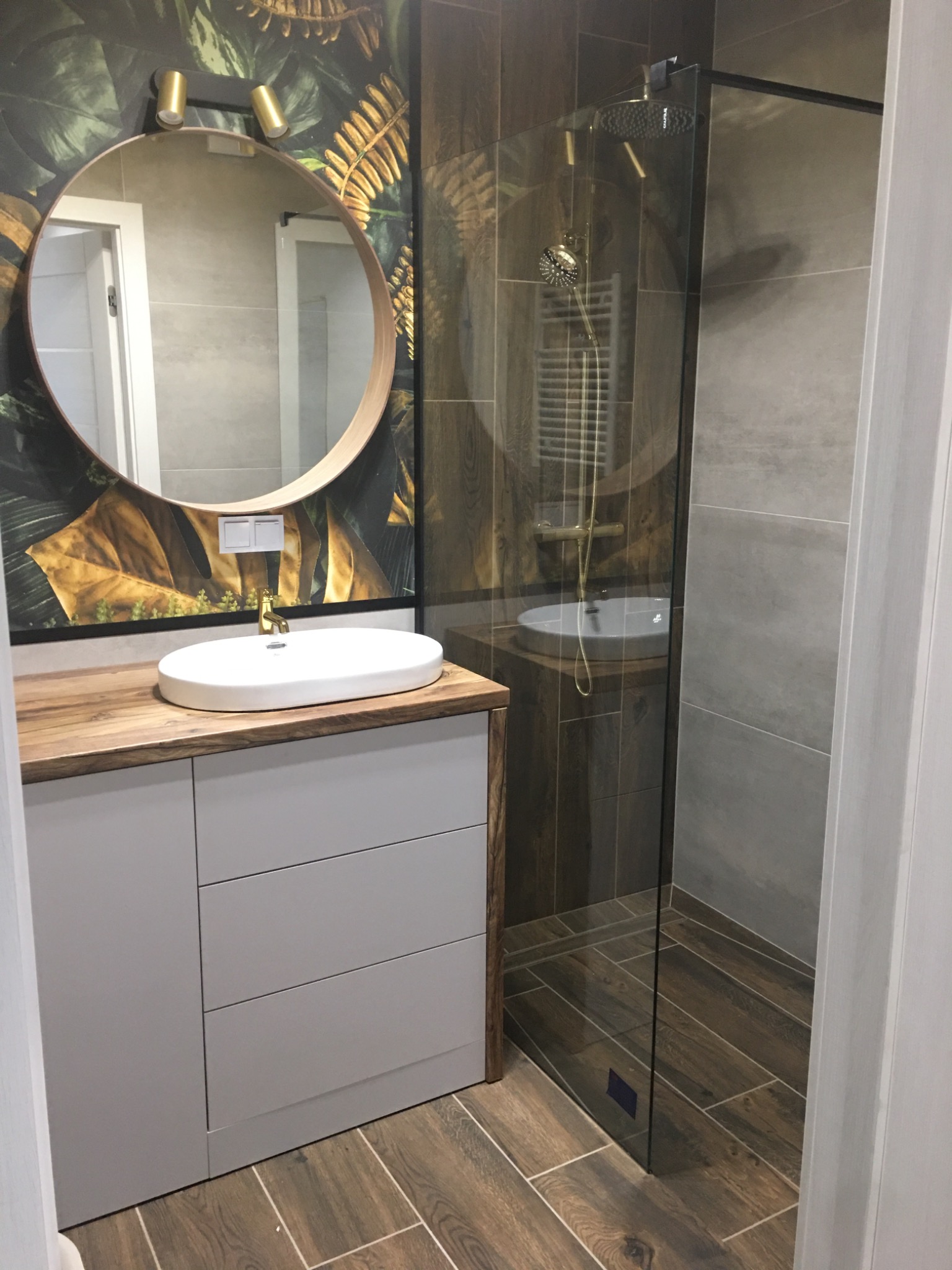 Nowoczesna łazienka z kabiną prysznicową typu walk-in, umywalką nablatową na drewnianym blacie i szafką w kolorze gołębim, okrągłe lustro podświetlone złotymi lampkami, ściana z motywem roślinnym.