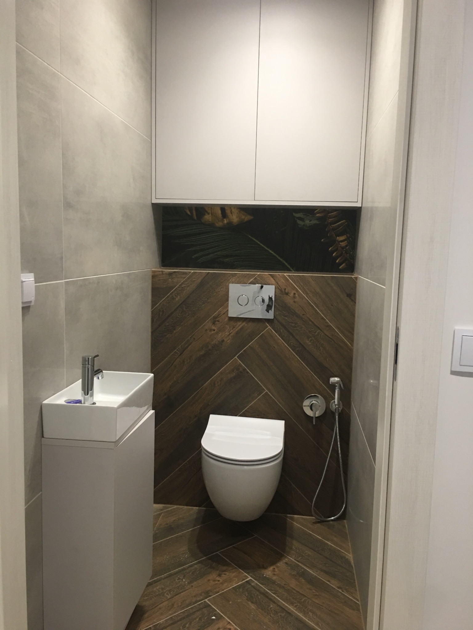 Nowoczesna łazienka z drewnopodobnymi płytkami ułożonymi w jodełkę, wiszącą toaletą i umywalką nablatową na szafce, oświetlona górnym światłem.